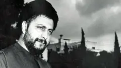 Foto en blanco y negro de Musa al-Sadr. Tiene barba y turbante negro y se ve pensativo. En el fondo se ven árboles y edificios