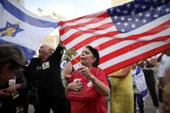 Uma mulher segura uma flor e parece tomada pela emoção, enquanto pessoas atrás dela acenam com bandeiras dos EUA e de Israel.