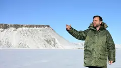 Джей Ди Вэнс в теплой куртке стоит на фоне заснеженной равнины и показывает на что-то рукой.