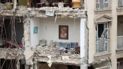 Bloco de apartamentos residenciais parcialmente destruído em Teerã, sem a parede externa e com um sofá azul em um cômodo repleto de escombros e um quadro ainda pendurado na parede