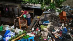 දිට්වා සුළි කුණාටුව හේතුවෙන් හානි වූ නිවසක් පිරිසිදු කරමින් සිටින කාන්තාවක්, ඇයගේ නිවාස ඉදිරිපිට කසළ ගොඩගසා තිබේ