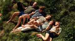 Vilijam na travi u kaki majici i plavom šorcu, pored njega je princ Džordž u plavoj majici i zelenom šorcu, Kejt se smeši pored njih u prugastoj majici i belim pantalonama, drži za ruku princa Luja koji nosi zelenu košulju i plavi karirani šorc, princeza Šarlot nosi crni šorc i bledozelenu majicu