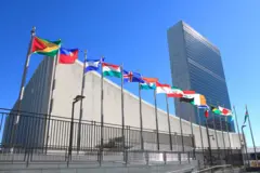 Sede da ONU em Nova York vista de fora, sob céu azul; é possível ver bandeiras de vários países