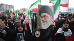 イラン国旗とハメネイ師の遺影を手にした黒衣の老若男女が、大勢集まっている。右手を挙げて叫ぶ人もいる。