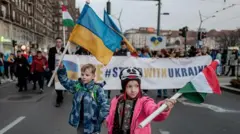 Двоє дітей йдуть попереду демонстрантів по вулиці у Будапешті, тримаючи у руках прапори України на Угорщини. Позаду них - люди з гаслами на підтримку України.