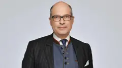 Um homem de óculos, paletó, colete, gravata e lenço branco no bolso do paletó.