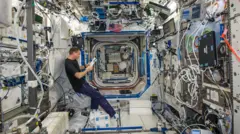 Wiseman flutua em gravidade zero dentro de um movimentado módulo da estação espacial, cercado por equipamentos, laptops e cabos emaranhados em todas as superfícies. Ele lê em um tablet enquanto bolsas de carga, experimentos e armários de armazenamento revestem as paredes e o teto. O laboratório estreito, semelhante a um túnel, se estende para os módulos adjacentes, iluminados por painéis brancos brilhantes. Cada espaço disponível está repleto de instrumentos, conferindo ao interior de alta tecnologia uma sensação densa e desordenada, ideal para pesquisas em microgravidade