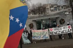 En primer plano, bandera venezolana, de fondo, personas manifestándose con pancartas donde se puede leer en inglés "Hands off Venezuela" - manos fuera de Venezuela.