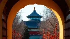 Uma vista através de um portal histórico de onde se vê edifícios de um templo em tons de azul, vermelho e dourado, com numerosas lanternas chinesas penduradas nas árvores sem folhas em primeiro plano.