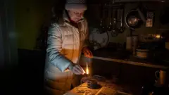 Женщина зажигает "окопную" свечу в темной кухне. 