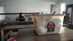 একটি টেবিলের ওপর ব্যালট ভর্তি একটি ভোটবাক্স, পেছনে একটি বেঞ্চের ওপর একই রকম আরেকটি বাক্স। আরও পেছনে একটি বেঞ্চে বসে আছেন একজন নারী