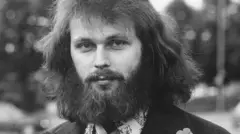 Fotografía en blanco y negro de Clark Olofsson en la década de 1970. Viste una camisa estampada con flores y un traje oscuro. Tiene una barba tupida y mira fijamente a la cámara.