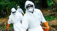 The image depicts a group of individuals dressed in full-body protective suits, complete with face masks, goggles, and bright orange gloves. Their attire suggests they are engaged in a high-risk operation, likely involving hazardous materials or infectious disease control. They are working outdoors in what appears to be a forested or rural environment, with greenery and natural terrain surrounding them. The scene conveys a sense of urgency and seriousness, as the protective gear is designed to minimise exposure to dangerous contaminants.