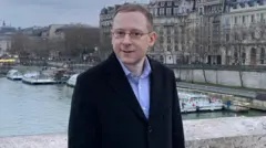 Un retrato de Nathan Cofnas de pie sobre un puente con agua y edificios al fondo.