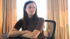 Claire Brosseau is photographed at her home in Toronto, Canada. She has dark brown hair and blue eyes. She is seated at a table with her arms crossed. She is wearing a brown short-sleeved turtle neck.