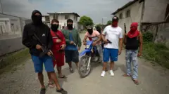 Sete jovens com balaclavas brandem armas automáticas em plena luz do dia, de frente para a câmera, dois deles em uma moto