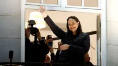 María Corina Machado saluda desde un balcón de un hotel en Oslo, Noruega. Tiene su mano extendida, sonríe y lleva el cabello negro hasta los hombros. viste un abrigo negro. Detrás hay varias personas con cámaras.