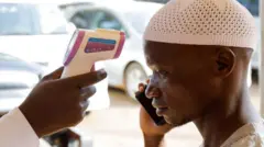 Una persona vestida de blanco aparece a la derecha de la imagen mientras utiliza un teléfono móvil y otra persona le mide la temperatura con un termómetro sin contacto; al fondo, varios coches aparecen desenfocados.
