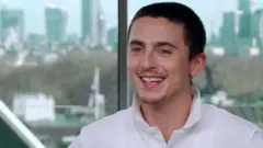 Timothée Chalamet sorri vestindo camisa pólo branca durante sua entrevista à BBC News, em frente a uma janela de vidro mostrando Londres ao fundo