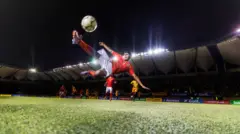 Kilku piłkarzy na boisku, na pierwszym planie jeden piłkarz ubrany w biało-czerwony strój kopie piłkę