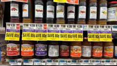 Potes de geleia Smucker e St Dalfour competem nas prateleiras de um supermercado com placas dizendo "Economize 80 centavos"