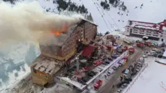 На фотографии видно, как горит одновременно крыша и некоторые помещения здания, находящиеся на разных этажах (огонь видно через окна). У здания треугольная крыша. Рядом парковка, на ней помимо обычных машин стоят пожарные.