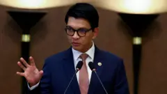 黒縁の眼鏡をかけ、青いスーツに赤いネクタイを締めた男性が、2本のマイクに向かって手を上げながら話している