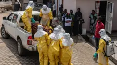 Profissionais de saúde usando equipamento de proteção individual (EPI) completo levantam o caixão de uma vítima na traseira de uma caminhonete em Bunia. Algumas pessoas com máscaras os observam do lado de fora de um hospital.