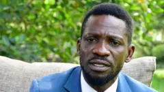 Bobi Wine yicaye, yambaye ikoti isa n'ubururu n'ishati yera. 