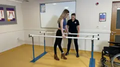 Megan está em pé segurando em duas barras. Uma mulher está ao lado dela e há uma cadeira de rodas na sala.