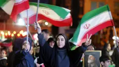 İran'da bayrak taşıyıp zafer işareti yapan, siyah çarşaflı iki kadın, arkada başka insanlar da var.