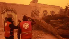 رجال إنقاذ تابعين للهلال الأحمر الليبي فرع الأبيار، تمكنوا من إخلاء عائلة بعد تساقط الأشجار على منزلها، بسبب الطقس والعواصف التي تضرب مدناً في شمال أفريقيا، ويظهر في الصورة رجلان من فريق الهلال الأحمر الليبي، يقفان أمام جذع شجرة محطم على الأرض، بينما تظهر السماء مصبوغة باللون الأحمر.