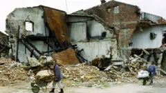Ethnic Albanians push wheelbarrows wey contain dia property in front of a ruined building, April 1999