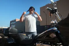 Daniel Overstreet, de 53 anos, veste uma camiseta branca e calça jeans enquanto coloca o capacete de motocicleta e se prepara para subir em sua Harley Davidson preta.