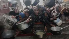 Um grupo de mulheres palestinas, vestidas com trajes muçulmanos e segurando panelas vazias, aguarda a distribuição de alimentos por uma instituição de caridade em meio ao bloqueio e aos ataques israelenses em Gaza.