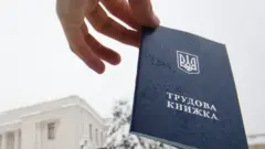 Чоловік високо тримає у руці трудову книжку. На задньому плані видніється будівля Верховної Ради.