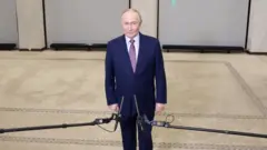 Путін у синьому діловому костюмі стоїть у кімнаті, перед ним тримають мікрофони 