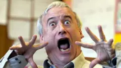 Роберт Манч строит смешную гримасу, подняв руки к лицу и открыв рот во время визита в школу Нельсона Манделы в Торонто в рамках празднования Дня семейной грамотности