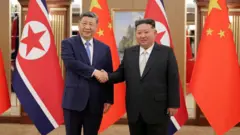 Xi Jinping e Kim Jong-un apertam as mãos. Os dois usam ternos. Ao fundo, há uma fileira de bandeiras da China e da Coreia do Norte
