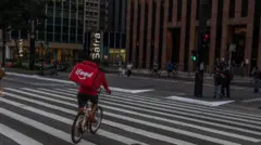 Um entregador anda de bicicleta enquanto faz uma entrega na Avenida Paulista, em São Paulo. Ele atravessa a faixa de pedestre e tem uma mochila vermelha com a marca iFood