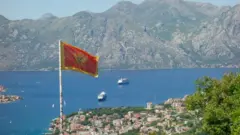 Yüksek bir yerden çekilen fotoğraf Karadağ bayrağı arkasında deniz manzarası ve dağları gösteriyor. 