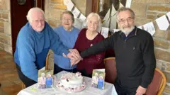 Dois casais celebrando 70 anos de casamento comemoram em frente a um bolo e cartões com retratos do rei Charles e da rainha Camilla