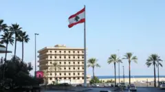 مركبات تسير بالقرب من العلم اللبناني الذي يرفرف في بيروت قرب الشاطئ، الصورة التقطت نهاراً.