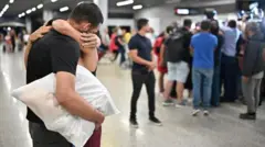 Duas pessoas se abraçando, com outras ao fundo, dentro de aeroporto