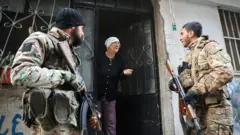 İki askeri kamuflaj kıyafetleriyle bir evin kapısının önünde duruyor, açık kapıdan dışarı doğru ileri yaşlı bir kadın, askerlerden sağdakiyle konuşuyor. 