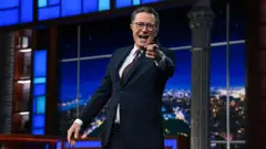 Stephen Colbert de pie en el set, con traje oscuro, señalando hacia la cámara.