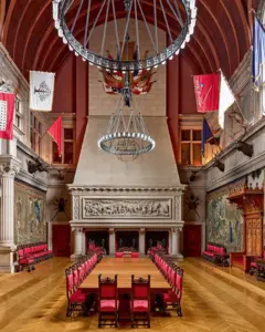 Sala com teto alto e tapeçaria nas paredes. No centro, uma longa mesa de madeira para banquetes, com cadeiras com estofamento vermelho, para cerca de 20 convidados.