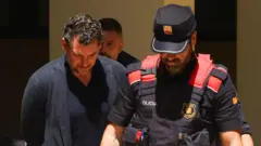 Muškarac u tamnom sakou pognute glave kojeg vodi policijski službenik