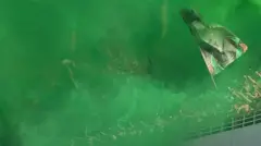 Ifoto igaragaza imbaga y'abafana bari mu kibuga kirimo umwotsi w'icyatsi kibisi uva ku bishashi by'umuriro w'ibyishimo, umwe mu bafana azunguza ibendera