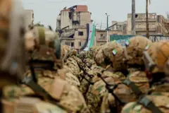 جنود مسلحون يتبعون للجيش السوري ويرتدون الزي العسكري يقفون في صفوف منتظمة خلال عرض عسكري في إدلب، ويظهرون من الخلف. وأمامهم العلم السوري بنجومه الحمراء الثلاثة.